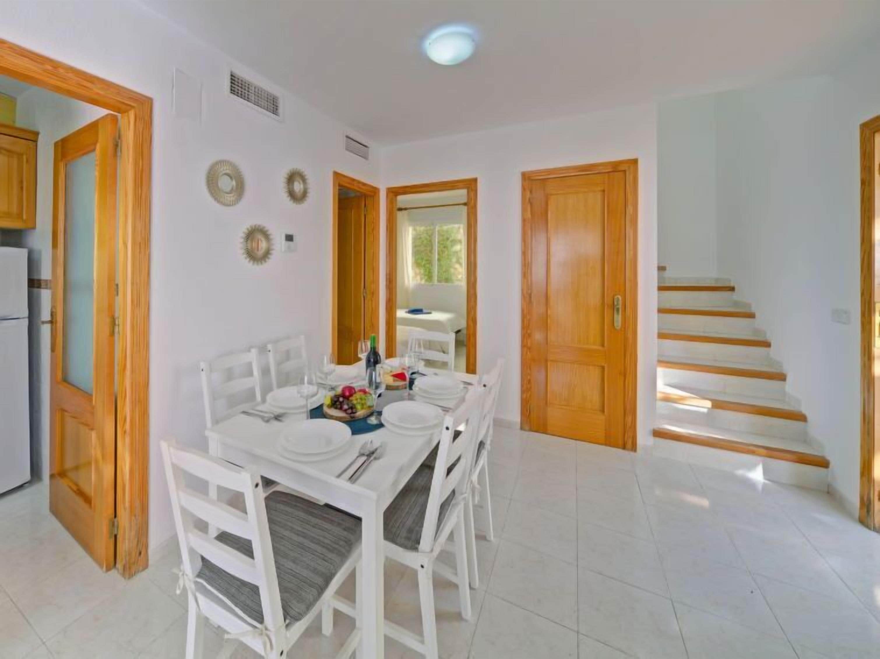 Ein Essbereich mit einem weißen Tisch, gedeckt für sechs Personen, umgeben von weißen Stühlen mit grauen Kissen. Holztüren und eine Treppe führen in einen weiteren Raum.
