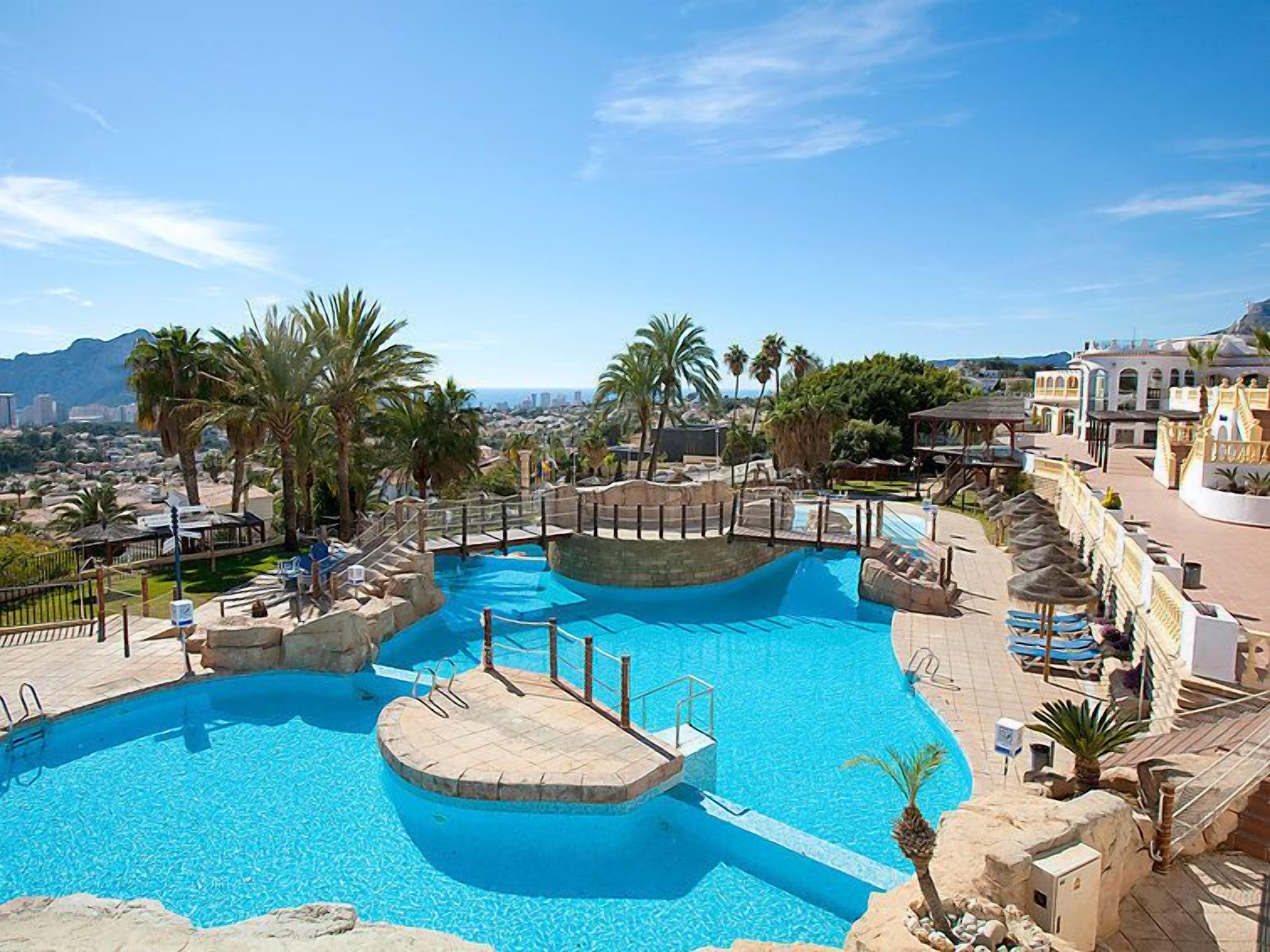 A large outdoor swimming pool with multiple levels and a surrounding deck area, set against a backdrop of palm trees and a cityscape.