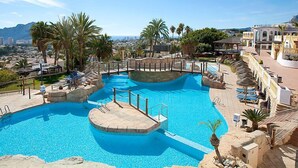 A large outdoor swimming pool with multiple levels and a surrounding deck area, set against a backdrop of palm trees and a cityscape.