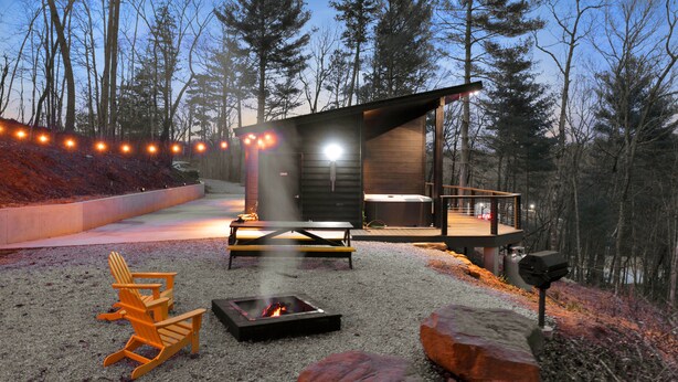 Eine moderne Blockhütte im Freien mit Feuerstelle, Sitzbereich und einer Terrasse mit Blick auf einen bewaldeten Bereich.