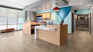 Una recepción con un mostrador de madera, una computadora y una lámpara colgante roja. Hay una máquina expendedora y una pared con un diseño azul y blanco.