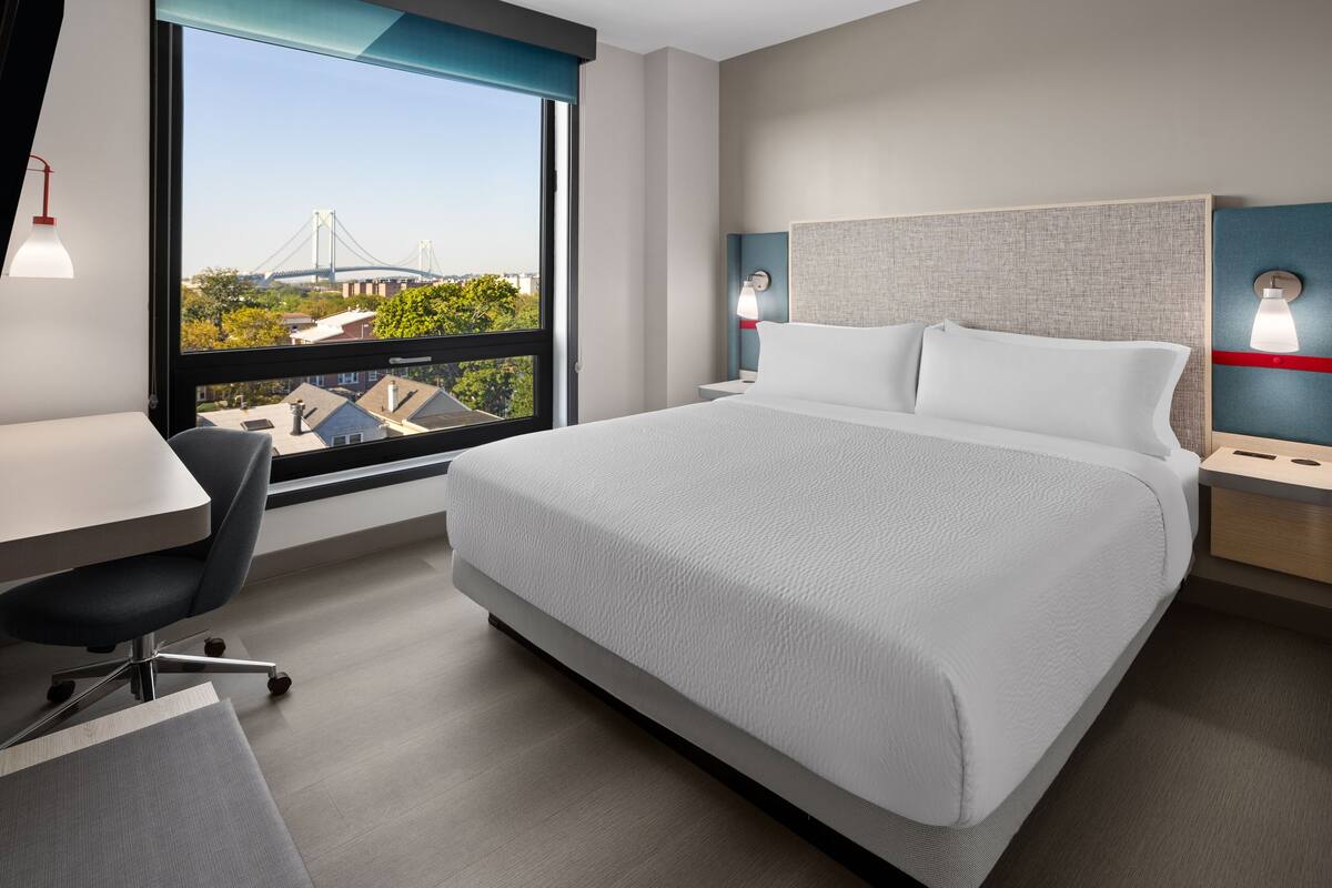 Une chambre d’hôtel avec un grand lit, un bureau et une chaise. La fenêtre offre une vue sur un pont et des immeubles.