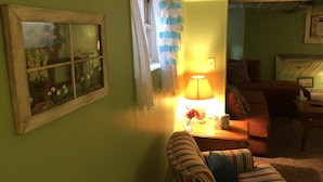 Un salon chaleureux avec un mur vert, une fenêtre donnant sur des fleurs, une lampe sur une table d’appoint et un canapé à rayures.