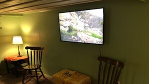 Une pièce avec un téléviseur fixé au mur, un fauteuil à bascule en bois, une petite table avec une lampe et une autre petite table recouverte d’une nappe jaune.