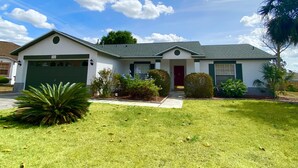A single-story house with a garage, a front door, and a small front yard with a palm tree and shrubs. - Modest Charmer W/ Private Pool (Clermont)
