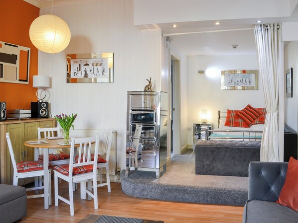 A modern living room with a dining area, a grey sofa, a white chair, a small table with a vase, and a bookshelf.