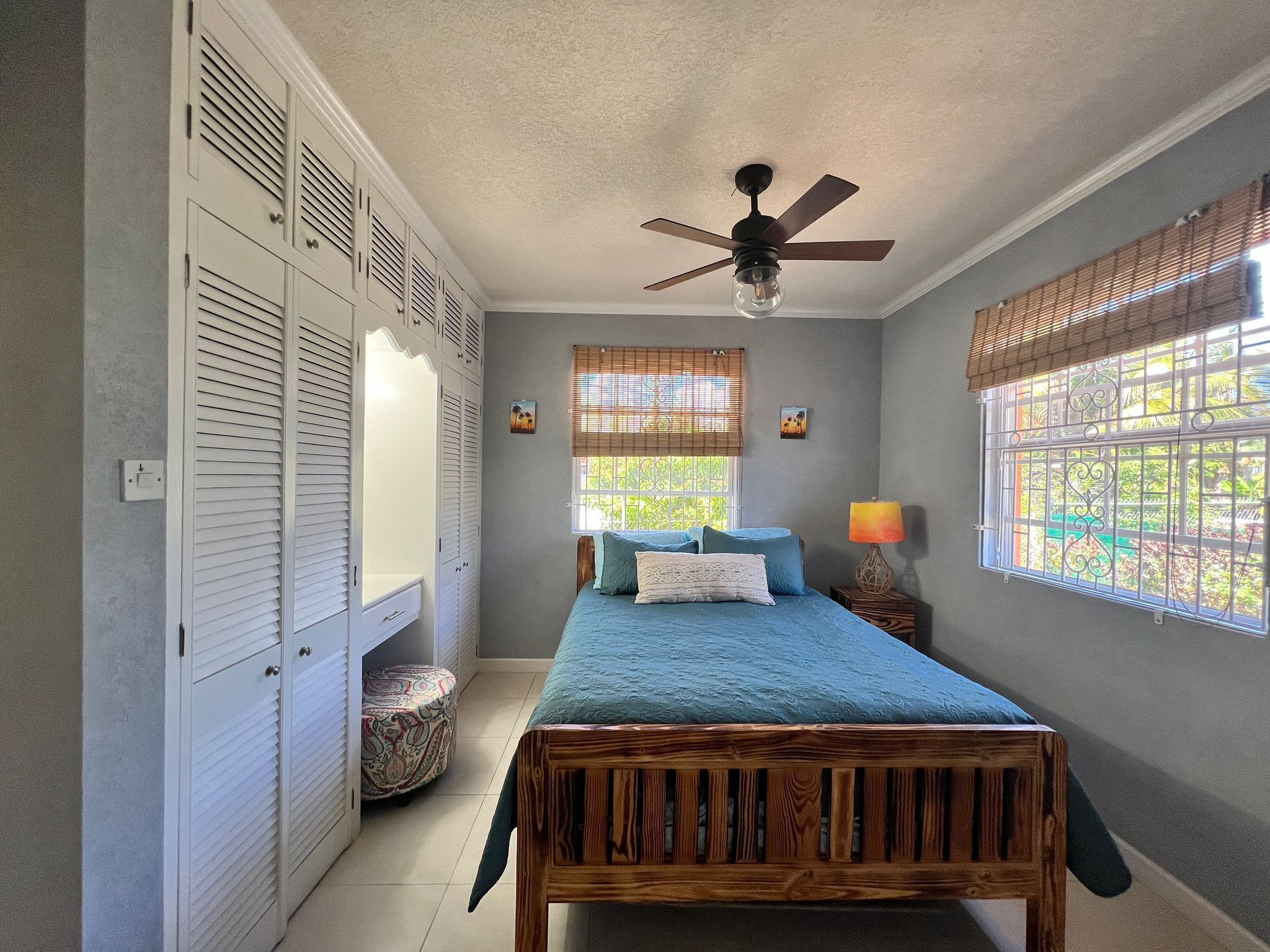 Une chambre à coucher comprenant un lit, un ventilateur de plafond, une fenêtre avec des stores, une lampe de chevet et une armoire avec des portes à persiennes.