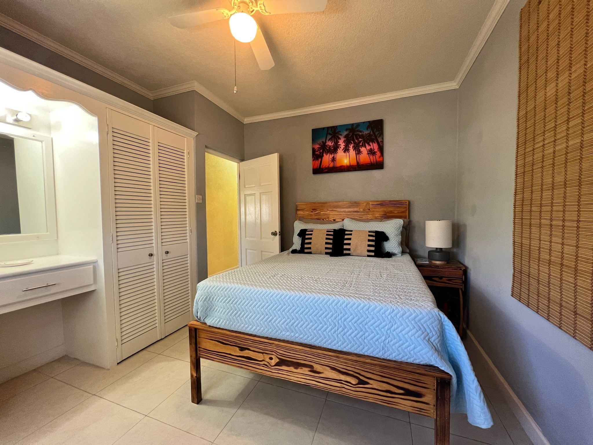A bedroom with a bed, a bedside table with a lamp, a wardrobe, a ceiling fan, and a painting on the wall.