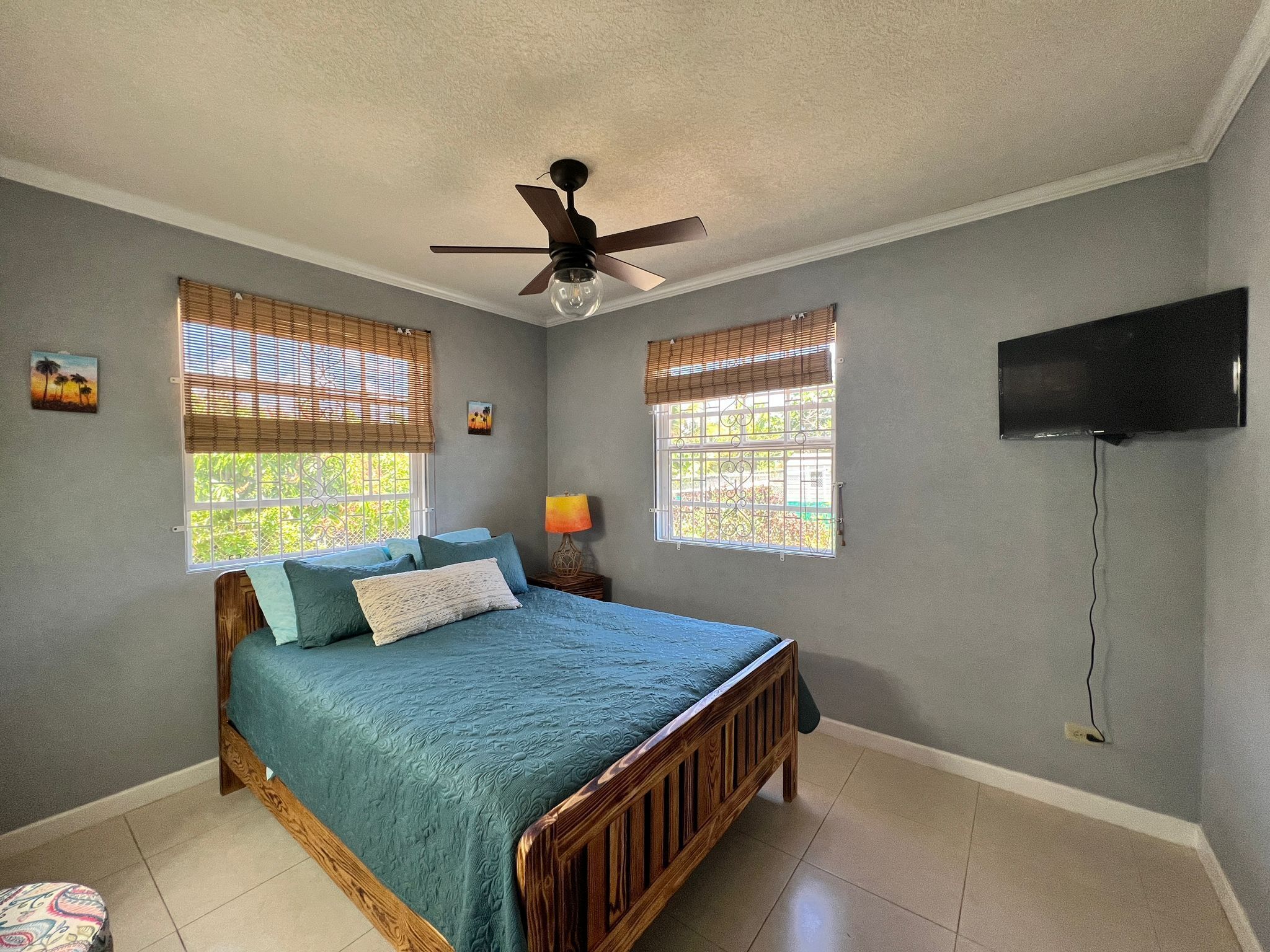 Un dormitorio con una cama, un ventilador de techo, dos ventanas con persianas, un televisor montado en la pared y una lámpara de noche.