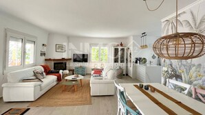 A modern living room with a white sofa, a coffee table, and a dining table with chairs. There is a TV mounted on the wall, a cabinet with glass doors, and a large window with curtains.
