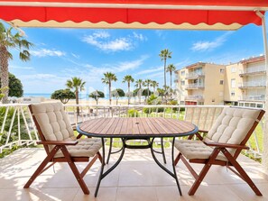 An outdoor patio with a round wooden table and two cushioned chairs, overlooking a beach and palm trees. - In 1st line of the beach, La Llosa area (Cambrils)
