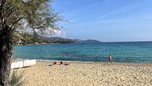 Ein Strand mit freiem Blick auf das Meer und Hügel in der Ferne. Liegestühle sind vorhanden und eine Person steht am Wasser.