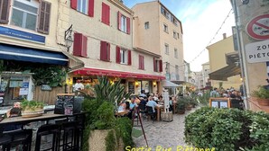Eine Kopfsteinpflasterstraße mit Außenbestuhlung eines Cafés, umgeben von Gebäuden mit roten Fensterläden und einem Schild, das ein Fahrradverbot anzeigt.