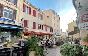 Eine Kopfsteinpflasterstraße mit Außenbestuhlung eines Cafés, umgeben von Gebäuden mit roten Fensterläden und einem Schild, das ein Fahrradverbot anzeigt.