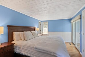 A bedroom with a wooden headboard, a large bed with white bedding, a nightstand with a lamp, and a window with blinds. - Contemporary Saltbox In New Seabury! (Mashpee)