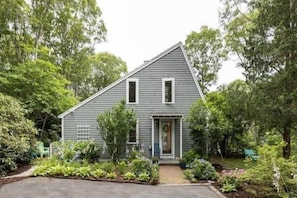 A single-story house with a grey exterior, a front porch, and a small garden with various plants and a bench. - Contemporary Saltbox In New Seabury! (Mashpee)