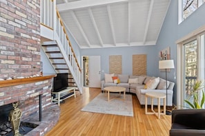A spacious living room with a brick fireplace, a wooden floor, a grey sofa, a coffee table, and a staircase leading to an upper level. - Contemporary Saltbox In New Seabury! (Mashpee)
