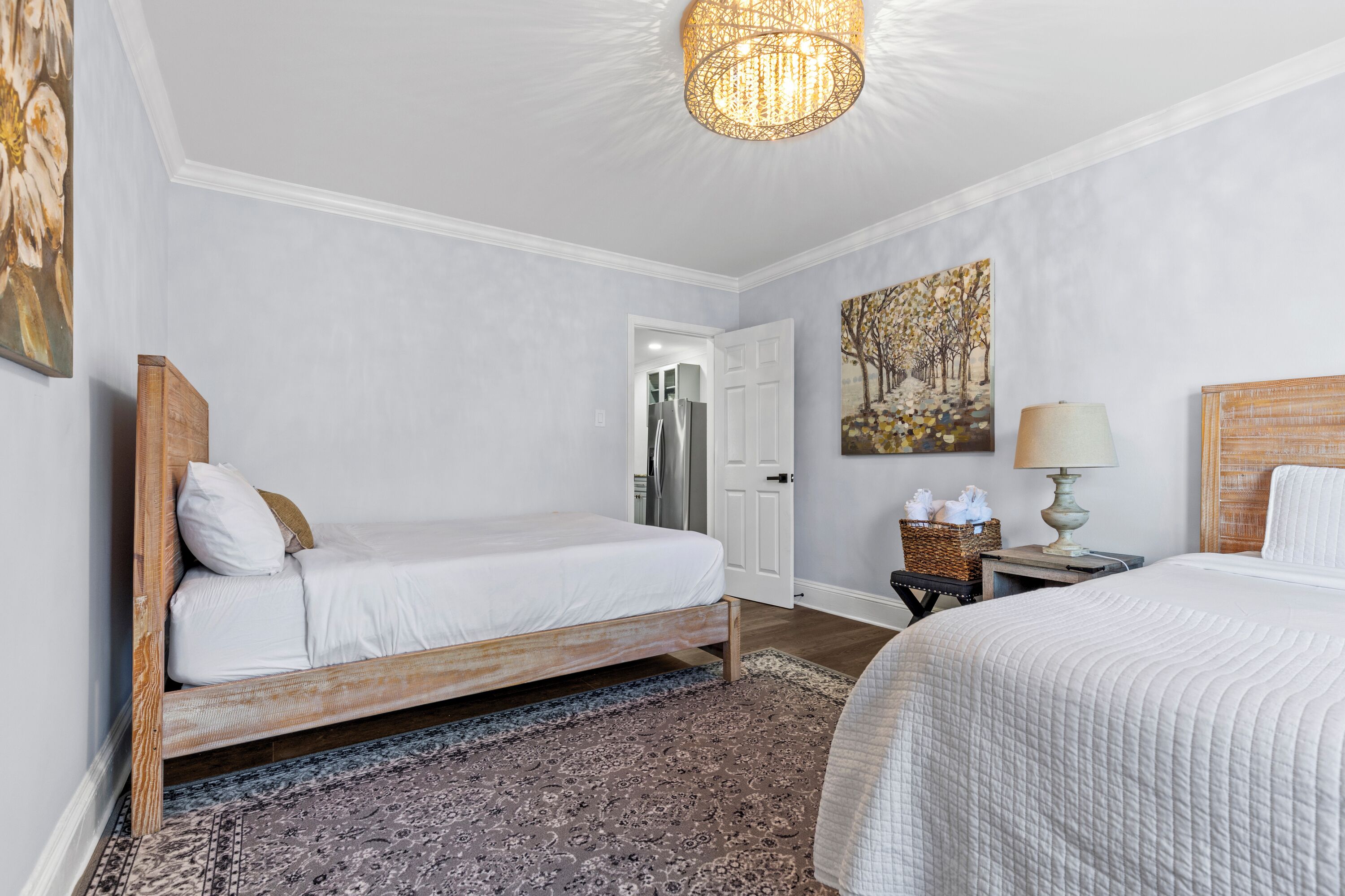 A bedroom with a wooden bed, a white bedside table with a lamp, a patterned rug, and a door leading to another room.