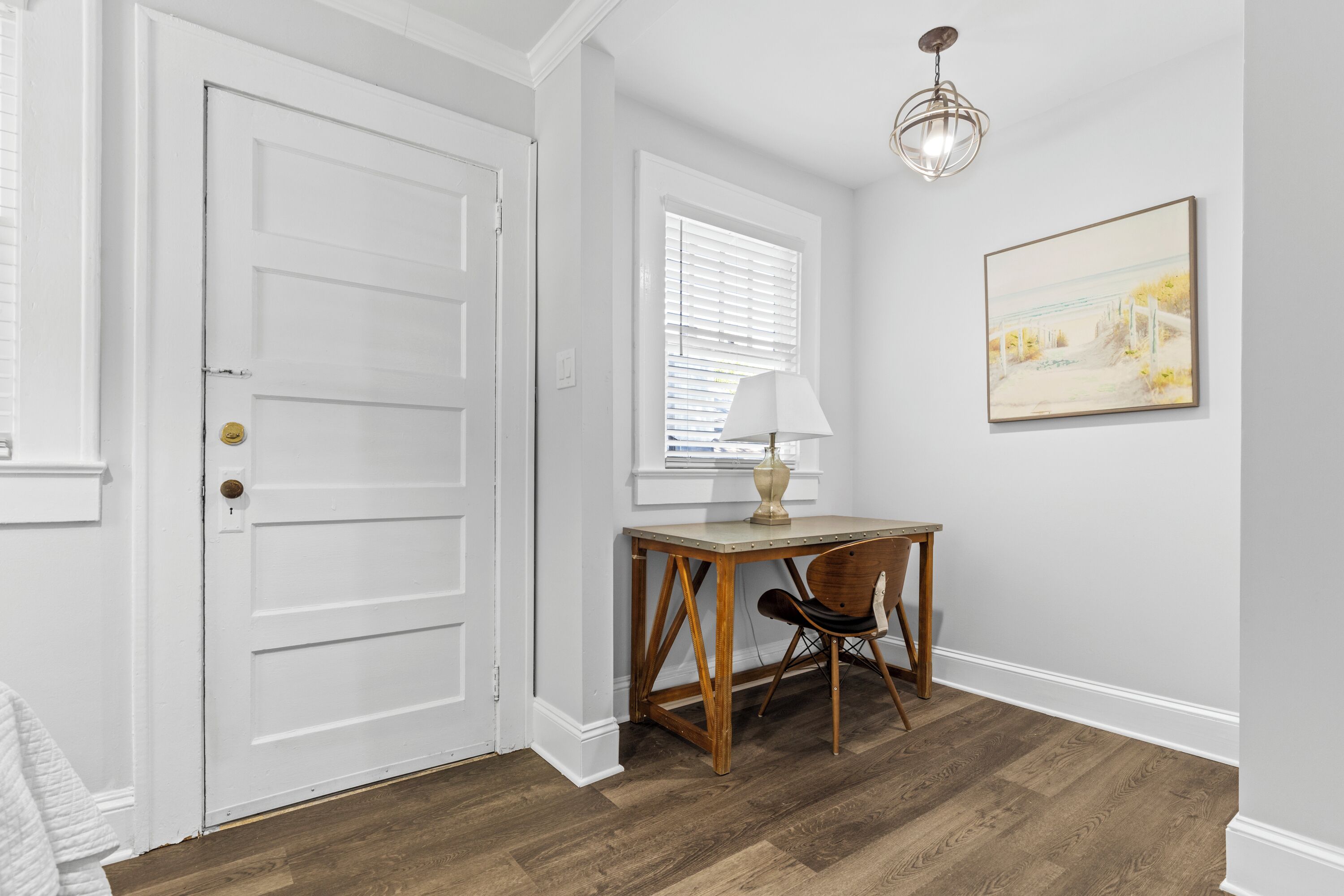 A room with a white door, a desk with a lamp, a chair, and a painting on the wall.
