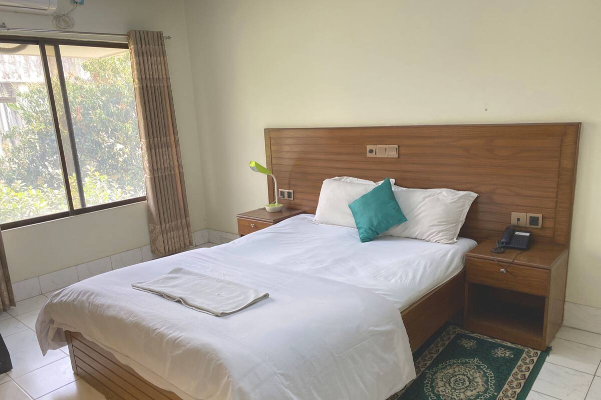 A neatly made bed with white linens, a teal pillow, and a bedside lamp. There is a small wooden nightstand with a phone and a power outlet. The room has a large window with curtains, a patterned rug, and a wooden headboard.