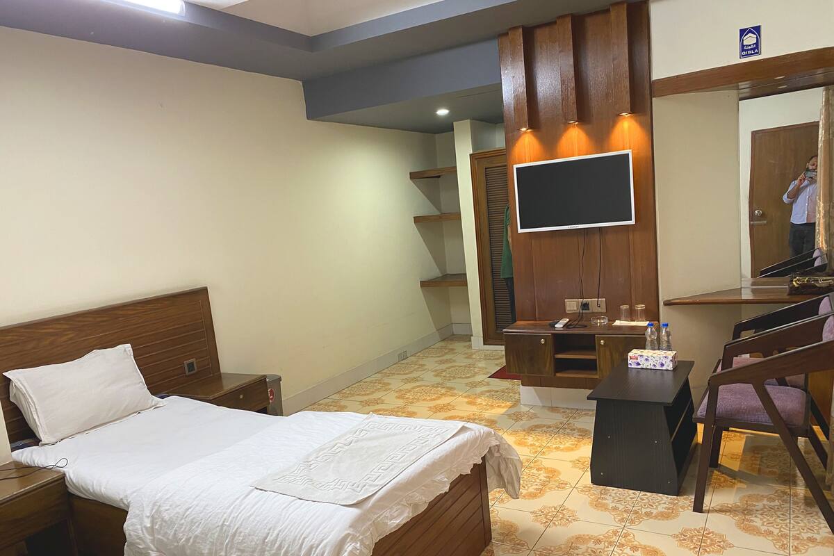 A hotel room with a bed, a desk with a flat-screen TV, a chair, and a small table with a box and bottles.