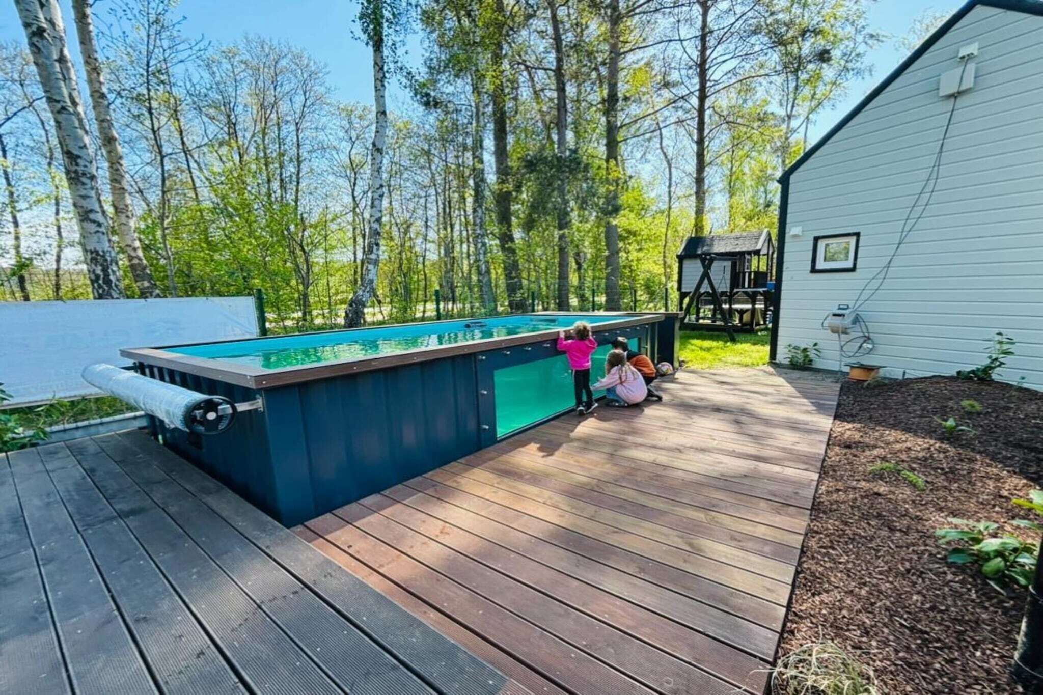 A pool area with a wooden deck, surrounded by trees and a white building.
