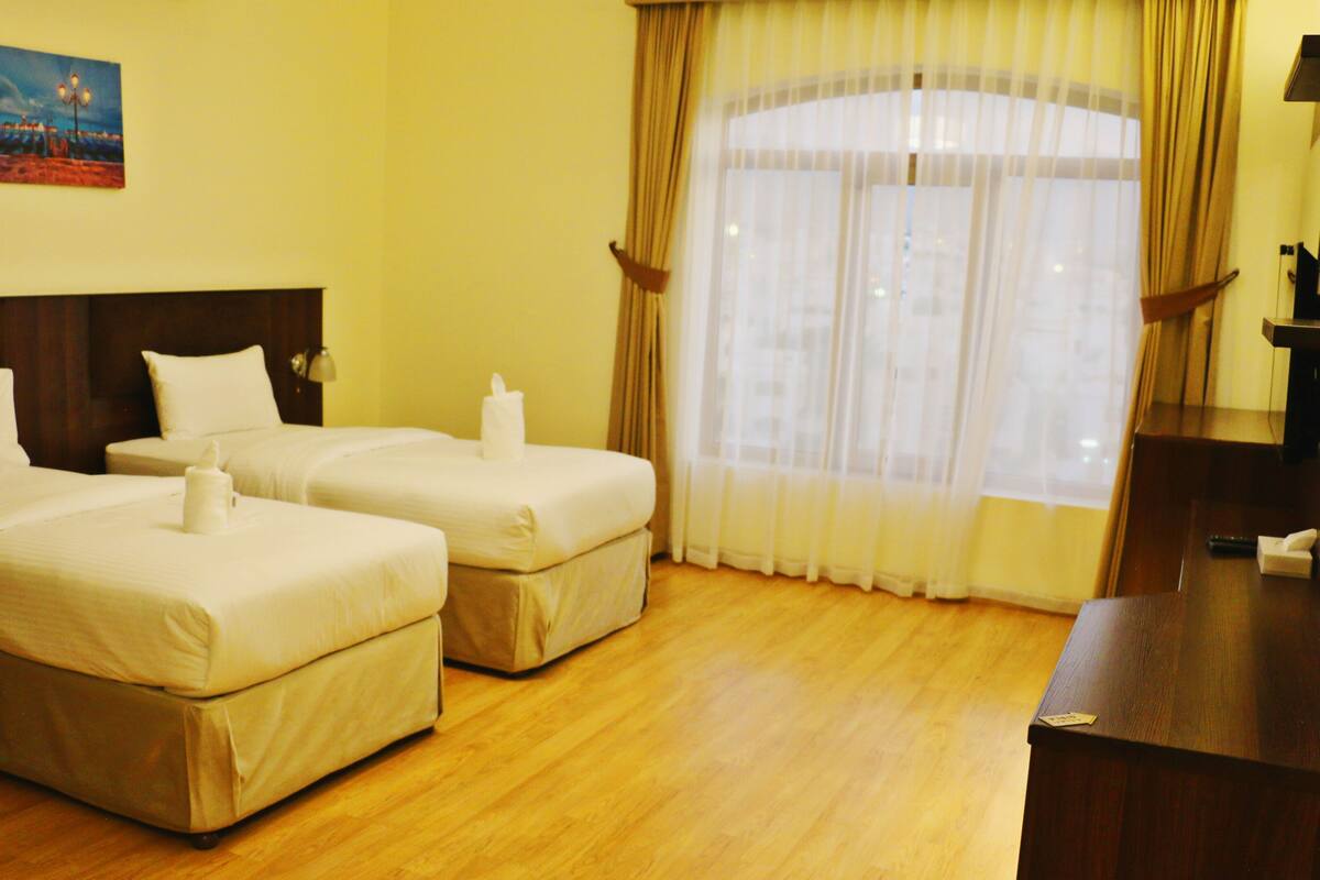 A hotel room with two beds, a wooden floor, a painting on the wall, and a window with curtains.
