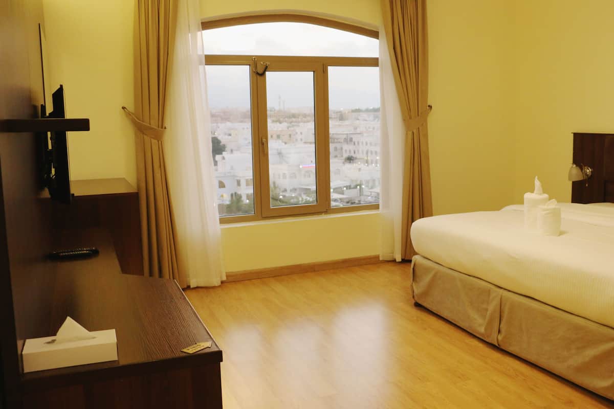 A hotel room with a large window, a bed with white linens, a wooden dresser, and a remote control on the dresser.