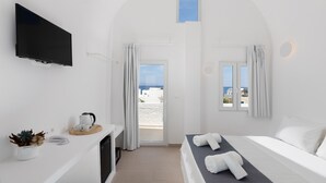 A hotel room with a bed, a TV, a small table with a kettle and cups, a plant, and a view of the beach. - VOCA HOTEL & SUITES (Santorini)