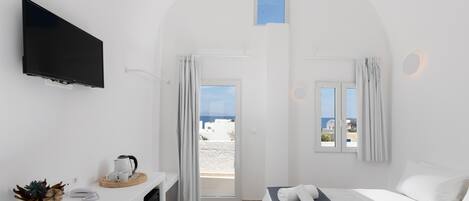A hotel room with a television, a small table with a kettle and cups, a bed with towels, and a view of the sea through the windows.