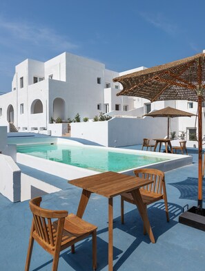 A poolside area with a wooden table and chairs, a pool, and a white building with arches. - VOCA HOTEL & SUITES (Santorini)