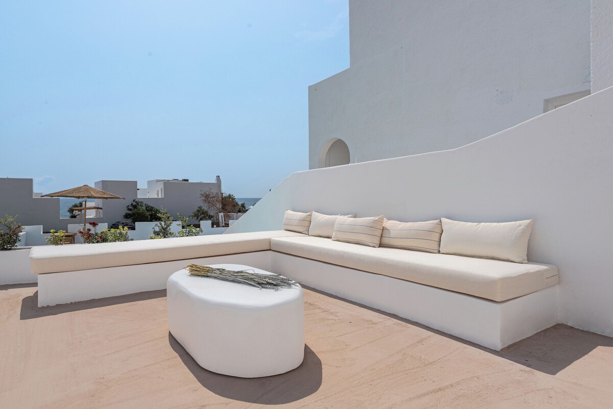 Un espacio al aire libre moderno con un sofá blanco y un reposapiés, una mesita y un paraguas decorativo.