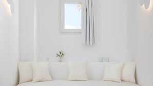 A minimalist white room with a sofa, a small window with curtains, and wall-mounted light fixtures. - VOCA HOTEL & SUITES (Santorini)