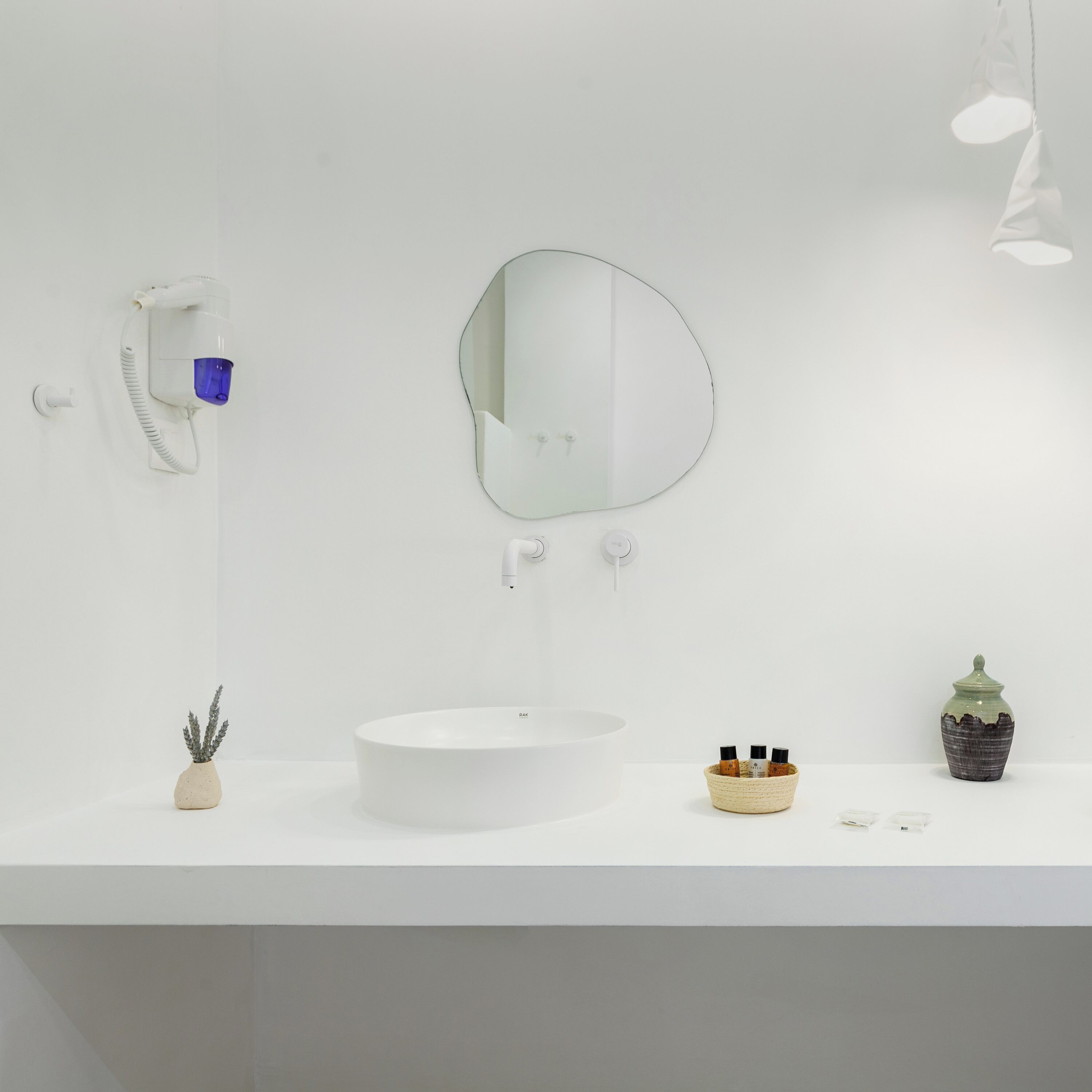 A minimalist bathroom with a white sink, a round mirror, a handheld showerhead, a small potted plant, and a decorative ceramic container.
