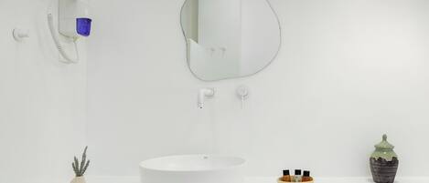 A minimalist bathroom with a white sink, a round mirror, a handheld showerhead, a small potted plant, and a decorative ceramic container.