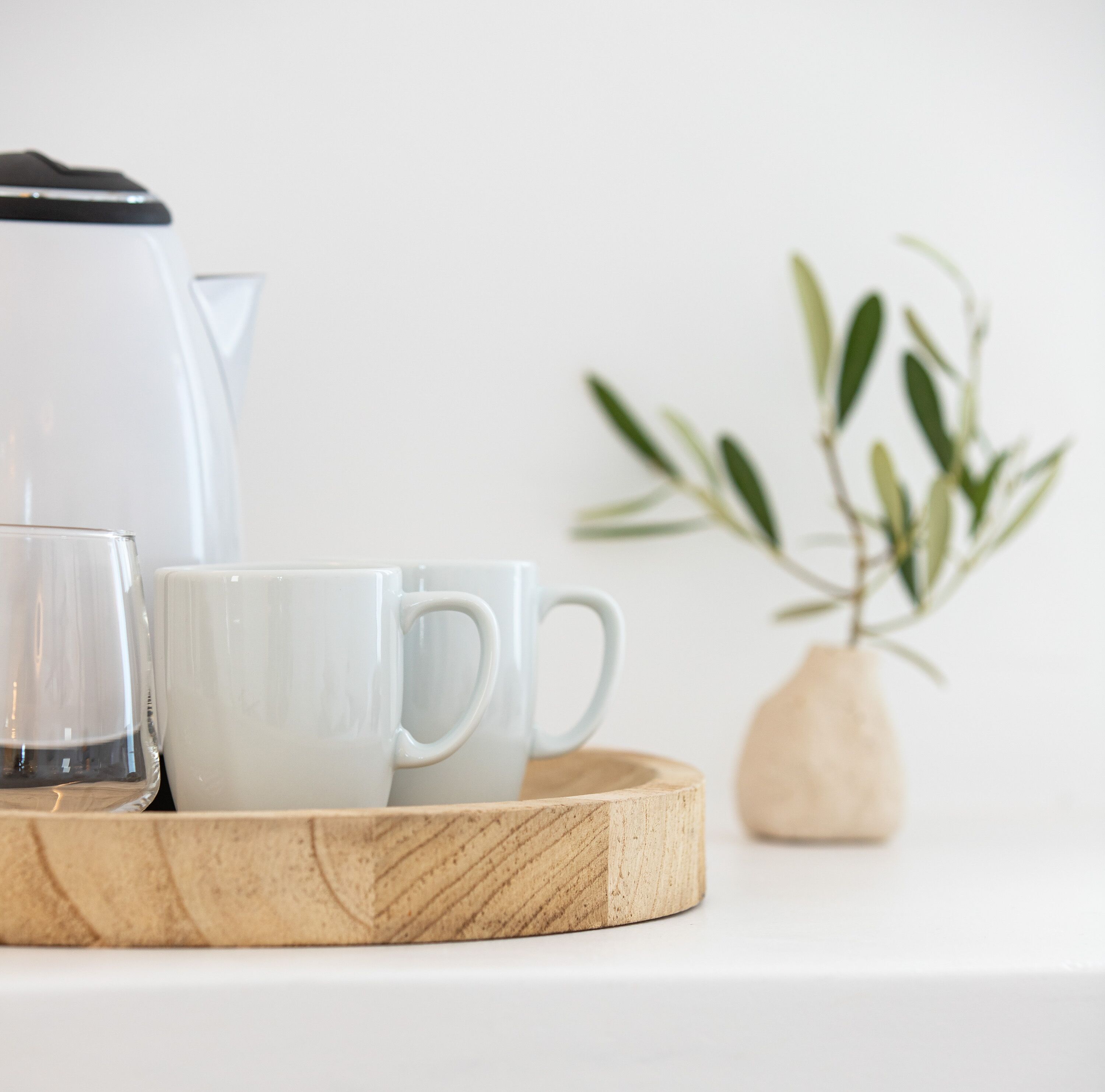 Una cafetera blanca, dos tazas blancas y una bandeja de madera sobre una superficie blanca.
