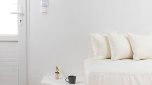 A minimalist room with a white sofa, a small table with a cup, and a wall-mounted light fixture. - VOCA HOTEL & SUITES (Santorini)