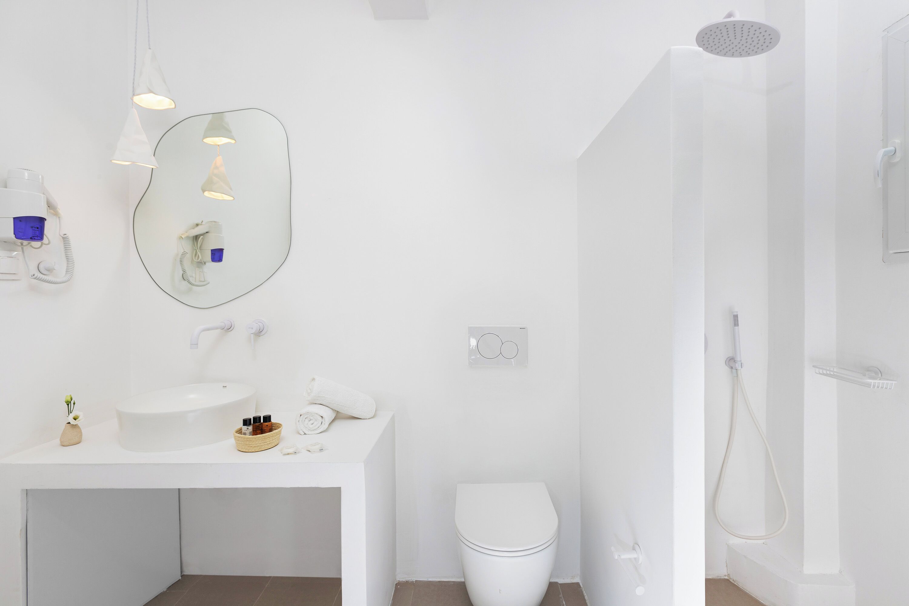 A modern bathroom with a white sink, mirror, and toilet. There is a shower with a handheld showerhead and a handheld dryer.