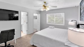 A bedroom with a bed, a desk, a chair, a ceiling fan, a window with blinds, and a wall-mounted TV.