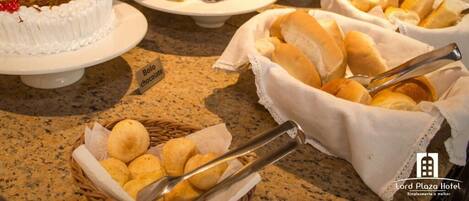 A buffet with various dishes including a cake labeled "Bolo chocolate," bread rolls, and other baked goods.