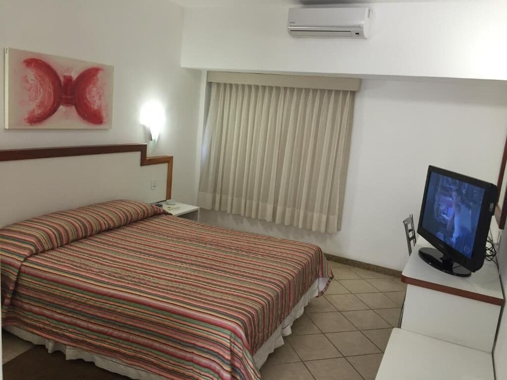 A hotel room with a bed, a television, a window with curtains, and an air conditioning unit.