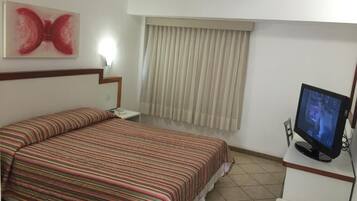 A hotel room with a bed, a television, a window with curtains, and an air conditioning unit.