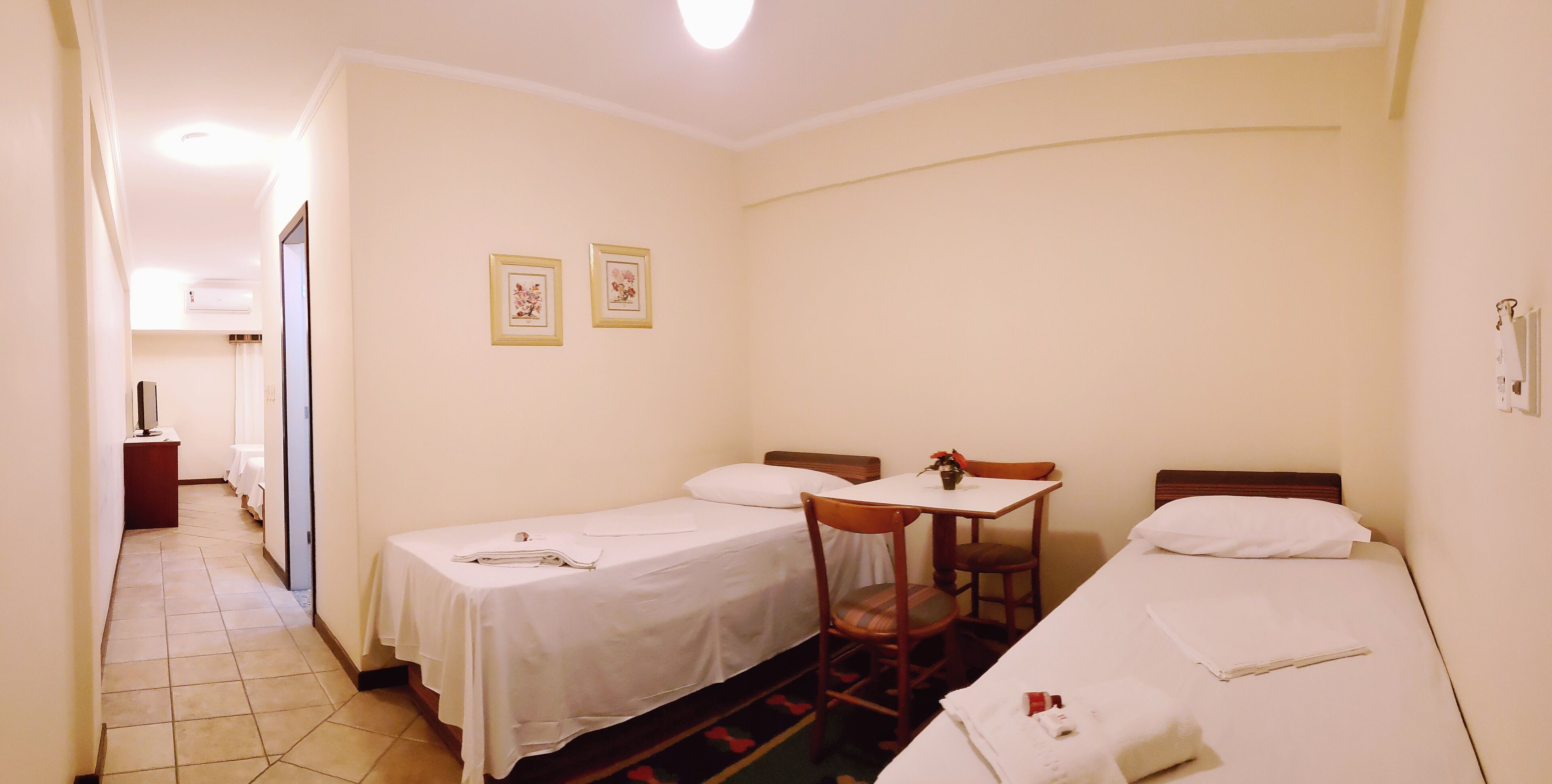A hotel room with two single beds, a small table with a chair, and a desk with a chair. There are two framed pictures on the wall.