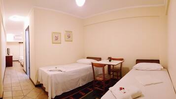 Quarto de hotel com duas camas de solteiro, uma pequena mesa com uma cadeira e uma escrivaninha com cadeira. Há duas quadros na parede.