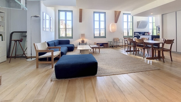 Un vaste espace de vie doté d’un haut plafond, de grandes fenêtres, d’une table à manger avec des chaises et d’un coin salon confortable avec un canapé et un pouf.