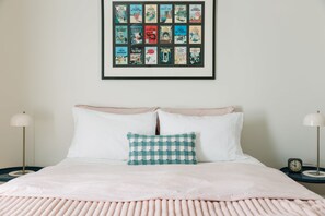 Un lit bien fait, orné d’un coussin à carreaux, une collection de bandes dessinées encadrées accrochée au mur, et des tables de chevet avec des lampes.