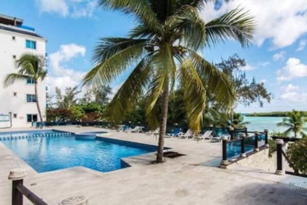 A pool area with clear blue water, surrounded by lounge chairs and palm trees, with a building in the background.