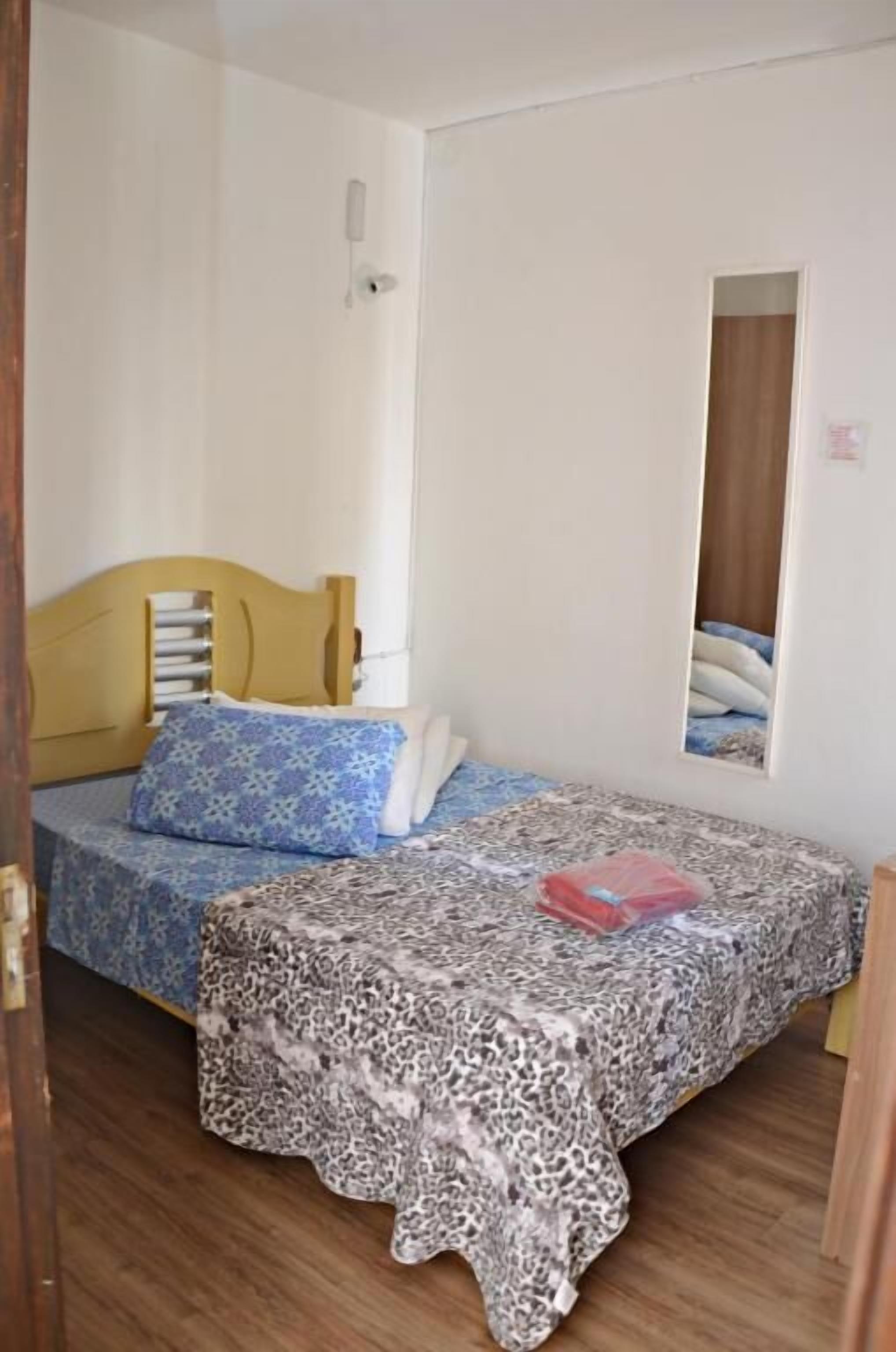 A small, single-bed room with a wooden headboard, a patterned bedspread, and a red object on the bed.