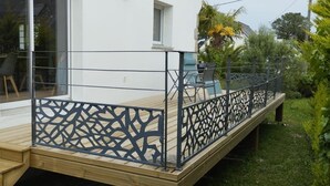 A wooden deck with a black lattice railing, a window, and a small table with chairs. - Maison golfe du morbihan (Locmariaquer)