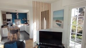 A modern kitchen with white cabinets, a wooden dining table, and a TV in a living area with a large window. - Maison golfe du morbihan (Locmariaquer)